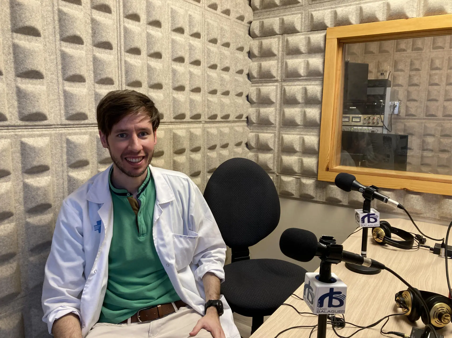 Psiquiatra integrativo en entrevista radial, vistiendo bata médica, sonriente ante un micrófono en un estudio con paneles acústicos, hablando sobre salud mental y enfoque holístico.
